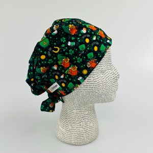 May include: Green, black, and orange surgical scrub hat with a pattern of leprechauns, shamrocks, gold coins, and the text "Lucky Day".