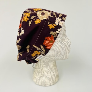 May include: A burgundy surgical scrub cap with a floral pattern in shades of orange, yellow, and cream. The cap is displayed on a mannequin head. The cap has a tag that says "Klinikaps".