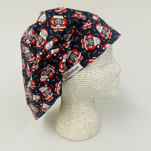 May include: A navy blue surgical scrub cap with a repeating pattern of red and white skeleton Santas and candy canes. The cap is made of fabric and has a tag that says "KimKaps".