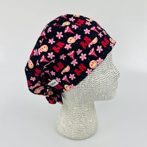 May include: Black scrub hat with a pink and red print of flowers, smiley faces, and the letters "RN" repeated.