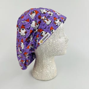 May include: A purple surgical scrub cap with a Halloween ghost print. The cap features white ghosts wearing orange hats and holding pumpkins, with black bats scattered across the fabric. The brand name "KimKaps" is visible on the side.