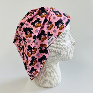 May include: A pink Halloween-themed cap with a repeating pattern of black bows, jack-o'-lanterns, and spiderwebs. The cap is worn on a mannequin head. The label on the cap reads "mkaps".