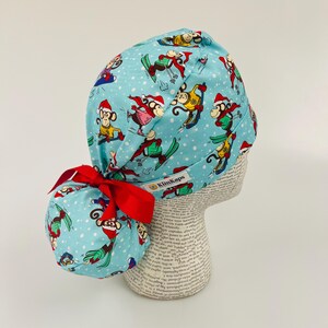 May include: A light blue surgical scrub cap with a repeating pattern of skiing monkeys wearing Santa hats. The cap has a red bow and the brand name "KimKaps" is visible on the side.