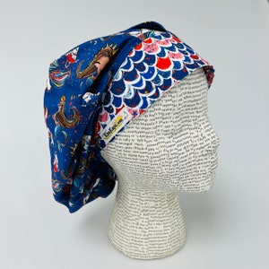 May include: A blue and white patterned surgical scrub hat with a mermaid design. The hat is reversible and has a second side with a red, white, and blue scale pattern. The hat has a small label that says "Kim Kaps.com".