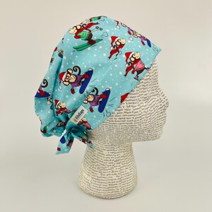 May include: A blue and white patterned surgical scrub hat with monkeys wearing Santa hats and skiing. The hat has a tie at the back and the text "KimKaps" is printed on the side.