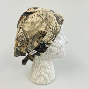 May include: A beige and brown patterned surgical scrub cap featuring a vintage-style print with bats, spiders, and trees. The cap is tied at the back and has a small label on the side.