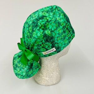 May include: Green scrub cap with a clover pattern and a matching green bow. The cap has a white tag that reads "KimKaps". The cap is designed to be worn on the head and is made of a soft, comfortable material.