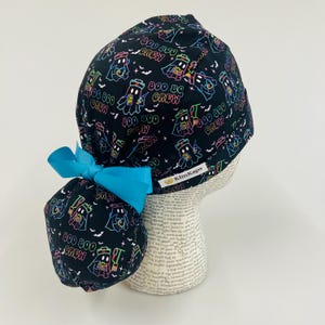 May include: Black scrub cap with a Halloween-themed pattern featuring ghosts, bats, and the text "BOO BOO CREW" in neon colors. A bright blue bow is attached to the back. The cap is designed for medical professionals.