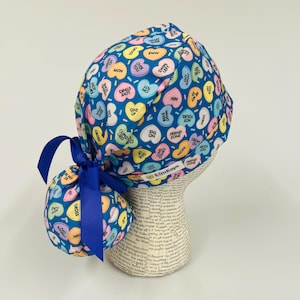 May include: A blue scrub cap and matching ponytail holder, both adorned with colorful candy heart graphics. The hearts feature phrases like "Love Stinks" and "Be Mine." A blue ribbon ties the ponytail holder.