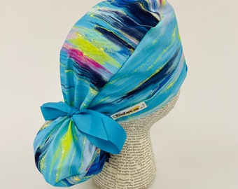 May include: A blue scrub cap with a colorful abstract design featuring streaks of yellow, pink, and navy. The cap has a matching blue bow and a small label that reads "KimKaps.com".