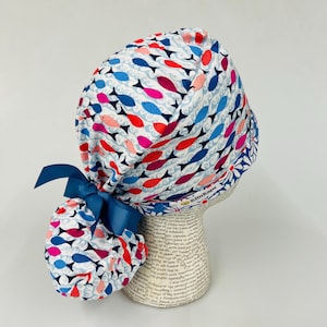 May include: A white surgical scrub cap with a repeating pattern of red, pink, and blue fish swimming on a wavy background. The cap has a dark blue bow and a floral patterned trim.