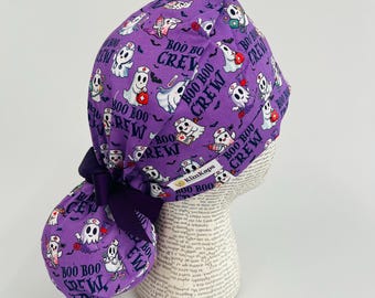 May include: A purple scrub cap with a Halloween theme. The cap features a pattern of cartoon ghosts dressed as nurses, bats, and the text "BOO BOO CREW". A dark purple bow is tied at the back. The brand name "KimKaps" is visible.