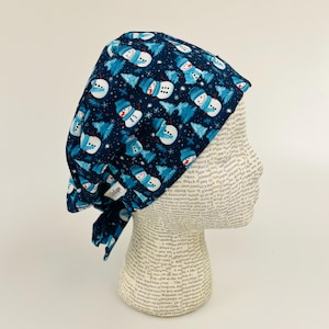 May include: A blue and white patterned surgical scrub hat with a snowman and pine tree design. The hat is tied on a mannequin head.