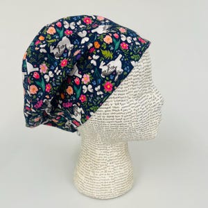May include: A navy blue surgical scrub cap featuring a floral and bunny pattern. The design includes white bunnies, pink and orange flowers, and white butterflies. The cap is made of a soft, flexible fabric.