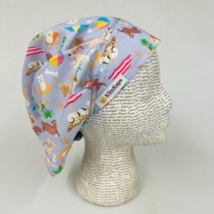 May include: A light blue surgical scrub cap with a beach-themed pattern featuring dogs, surfboards, beach umbrellas, and palm trees. The cap has a tag that reads "KimKaps".