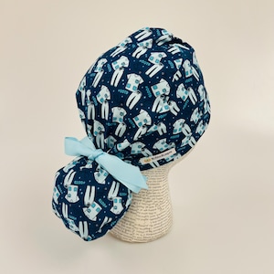 May include: A navy blue scrub cap with a light blue bow. The cap is patterned with white and light blue scrubs and the text "#OOTD". The cap is displayed on a mannequin head.