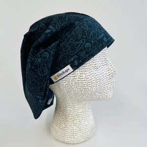 May include: A dark teal scrub cap with a Thanksgiving-themed pattern of pumpkins, turkeys, and acorns. The cap has the word "THANKFUL" printed on it. A small tag with the brand name "KimKaps" is visible.