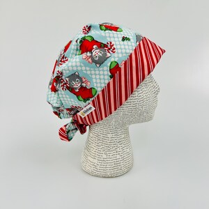 May include: A blue and red surgical scrub hat with a Christmas cat print. The hat has a red and white striped trim and a tie at the back.