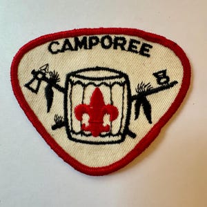 Może przedstawiać: Trójkątna naszywka z kremowym tłem i czerwoną obwódką. U góry napis "CAMPOREE". Czarny bęben z czerwoną lilią w środku. Czarne postacie flankują bęben, trzymając przedmioty.