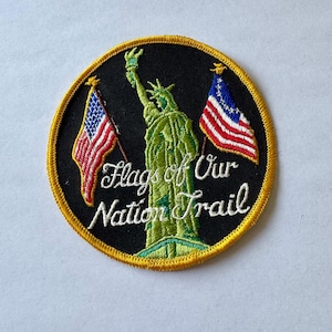 May include: A round, black embroidered patch with a gold border. The patch features a green Statue of Liberty with two American flags on either side. The text "Flags of Our Nation Trail" is embroidered in white on the patch.