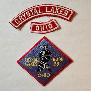 May include: Three embroidered patches. The top two are red with white text: "CRYSTAL LAKES" and "OHIO". The bottom patch is blue with a red border, featuring a black figure and text: "CRYSTAL LAKES", "TROOP 28", and "OHIO" in yellow.