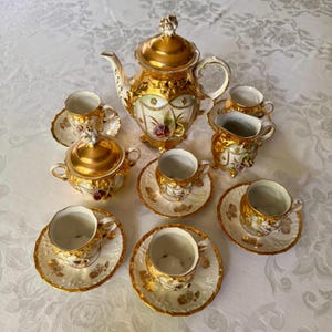 May include: A porcelain tea set featuring a teapot, a lidded sugar bowl, a creamer, and four teacups with matching saucers. Each piece is decorated with ornate gold trim, raised relief details, and painted floral accents on a white base.