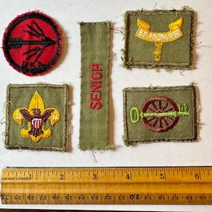 Op de afbeelding: Een verzameling vintage Boy Scout-patches. De patches hebben verschillende ontwerpen, waaronder een pijl, een adelaar, een sleutel en de woorden "BE PREPARED" en "SENIOR". Een houten liniaal met centimeter metingen staat onderaan.