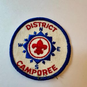 Może przedstawiać: Okrągła naszywka z niebieską obwódką i kremowym środkiem. Naszywka zawiera czerwone słowa "DISTRICT CAMPOREE", otaczające niebieski wzór róży wiatrów. W środku znajduje się czerwony fleur-de-lis.