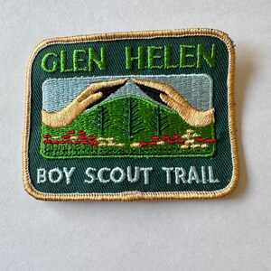 May include: Green embroidered patch with gold trim, featuring the text "Glen Helen Boy Scout Trail". The patch depicts a mountain scene with two hands reaching up towards the peak.