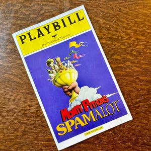Può includere: Un programma di sala per Monty Python's Spamalot, con una copertina gialla e viola. La parte superiore recita "PLAYBILL" in grandi lettere gialle. La copertina raffigura un calice d'oro tenuto da una mano.