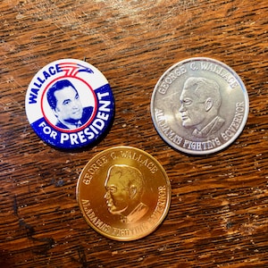 May include: Three campaign badges featuring George C. Wallace. The first badge is blue and white with the text "Wallace for President". The second badge is silver with the text "George C. Wallace Alabama's Fighting Governor". The third badge is gold with the text "George C. Wallace Alabama's Fighting Governor".