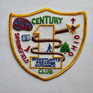 May include: Embroidered patch with a shield shape, featuring text and graphics. The patch includes the words "CENTURY," "SPRINGFIELD," and "OHIO." Graphics include a person, a bird, a tree, and an eagle. The patch is bordered with gold thread.