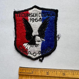 Może przedstawiać: Haftowana naszywka w kształcie tarczy, z czerwoną, białą i niebieską kolorystyką. Naszywka przedstawia orła z rozpostartymi skrzydłami, tekst "TECUMSEH COUNCIL 1964" i "CAMP-O-RAMA". Naszywka ma około 7.6 cm szerokości.