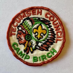 Può includere: Una toppa ricamata bianca con un bordo marrone. La toppa presenta la testa di un capo nativo americano con un copricapo verde, bianco e arancione. Un giglio giallo si trova al centro della toppa. Il testo "Tecumseh Council Camp Birch" è intorno al bordo.