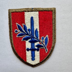 May include: Embroidered patch with a shield shape. The patch features a red background with a vertical white stripe. A blue sword and olive branch are centered on the white stripe. The patch is bordered with a brown stitched edge.