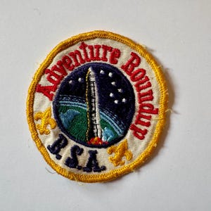 May include: Embroidered patch with a circular design. The patch features a rocket launching into space, surrounded by the words "Adventure Roundup" and "BSA". The patch has a yellow border and includes a blue, green, and white color scheme.