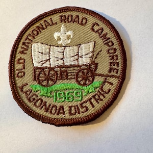 Pode incluir: Um remendo bordado marrom e bege com fundo verde. O remendo apresenta uma carroça com uma lona branca e um símbolo de escoteiro. O texto no remendo diz "Old National Road Camporee Lagonda District 1969".