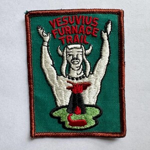 Peut inclure: Écusson brodé avec un fond vert et une bordure marron. Le patch représente une figure blanche avec les bras levés, des cornes et un volcan en éruption de lave rouge. Le texte "VESUVIUS FURNACE TRAIL" est arqué en haut en rouge.