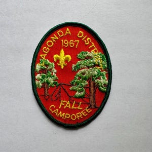 Może przedstawiać: Okrągła naszywka z czerwonym tłem i zieloną obwódką. Naszywka zawiera haftowany napis "AGONDA DISTRICT 1967 FALL CAMPOREE". Złoty fleur-de-lis i dwa drzewa są również wyhaftowane na naszywce.