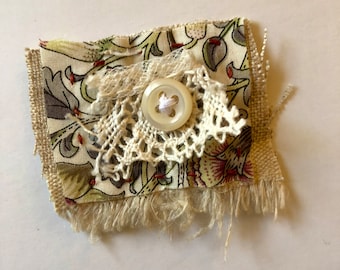 May include: A small piece of fabric with a floral pattern, a white button, and a lace overlay. The fabric is frayed around the edges.