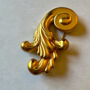May include: A gold-colored brooch with a stylized leaf and scroll design. The brooch has a pin on the back for attachment. The design features a detailed, textured surface and a bright, shiny finish. This vintage-style accessory is suitable for adding a touch of elegance.