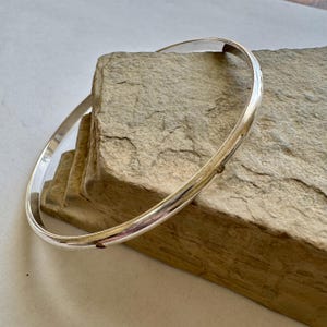 May include: A shiny, silver bangle bracelet. The bracelet is round and smooth, reflecting light. The jewelry is resting on a textured, light brown stone surface. The bangle is a simple, elegant piece of jewelry.