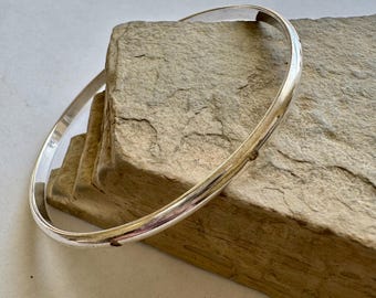 Op de afbeelding: Een glanzende, zilveren armband. De ronde armband is glad en reflecteert licht. Het ligt op een getextureerd, lichtbruin stenen oppervlak. De armband is een eenvoudig, elegant sieraad.