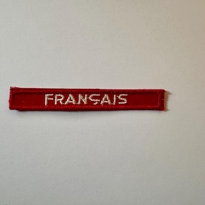 Può includere: Una toppa rettangolare rossa con la scritta "FRANÇAIS" ricamata in bianco. La toppa è realizzata in tessuto strutturato ed è probabilmente progettata per essere cucita su abiti o altri oggetti.