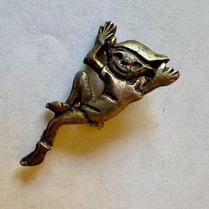 May include: A vintage metal pin in the shape of a whimsical elf or pixie. The pin features a smiling face, pointed ears, and outstretched arms and legs. The metal has a tarnished, antique gold finish.