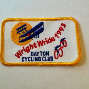 Pode incluir: Remendo bordado com borda amarela e fundo branco. O remendo apresenta um biplano azul em frente a um sol amarelo, com o texto "Wright Wride 1993" em vermelho. Abaixo, lê-se "Dayton Cycling Club" em azul.