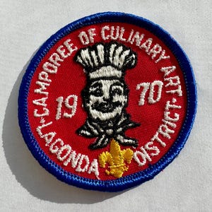 Może przedstawiać: Naszywka haftowana na czerwono i niebiesko z kreskówkowym kucharzem i tekstem "1970 Lagonda Camporee of Culinary Arts District".