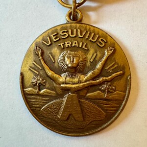 May include: A bronze-colored medal with the words "VESUVIUS TRAIL" arched across the top. The medal features a stylized figure with outstretched arms, a mountain, and two trees. The letter "A" is at the bottom.