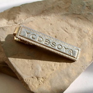 Op de afbeelding: Zilverkleurige stropdasklem met de gegraveerde tekst "YCDBSOYA". De rechthoekige klem heeft een touwachtige rand en een goudkleurige sluiting. Het ligt op een lichtbruin, getextureerd stenen oppervlak.