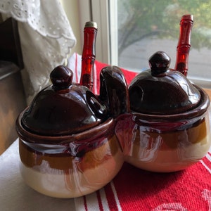 May include: A brown and white ceramic double condiment set with two red and white glass stoppers. The set has two lids and two small bowls.